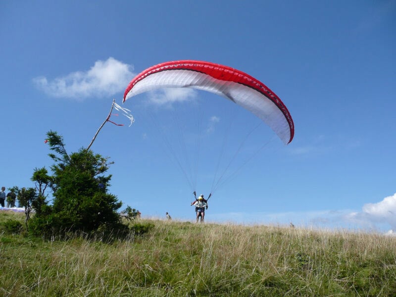 Parapentiste en pleine phase de décollage sur une colline herbeuse, avec une voile rouge et blanche se déployant sous un ciel bleu dégagé.