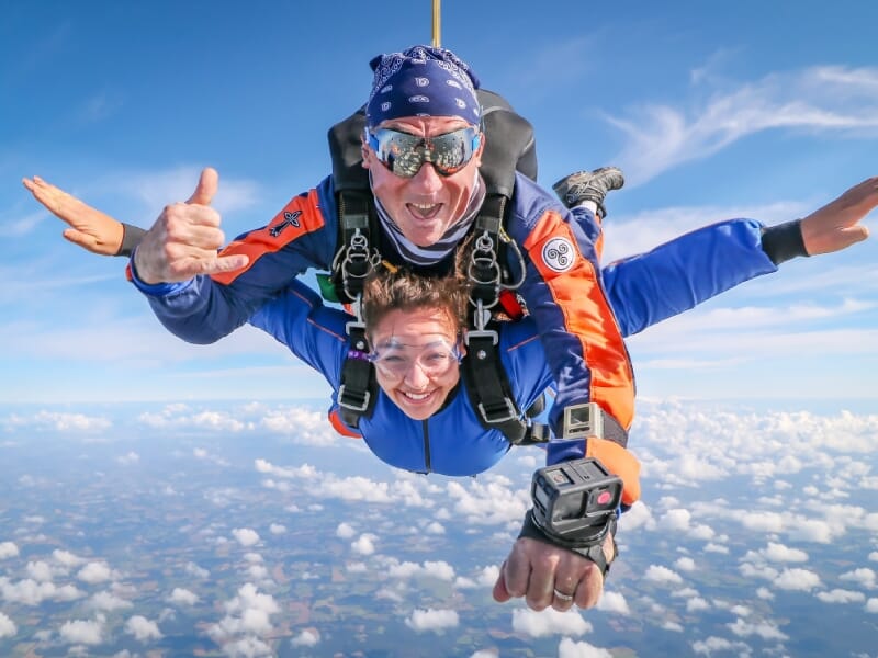 Tandem parachutistes en chute libre, homme et femme souriants avec pouces levés, tenues de saut bleue et orange, lunettes de protection, au-dessus des nuages blancs, ciel clair.