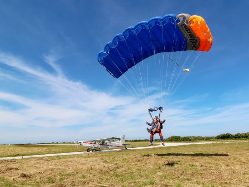 Atterrissage de parachutiste en tandem, parachutiste et passager touchant le sol, parachute bleu et orange au-dessus, avion blanc sur la piste en arrière-plan, ciel bleu, herbe verte