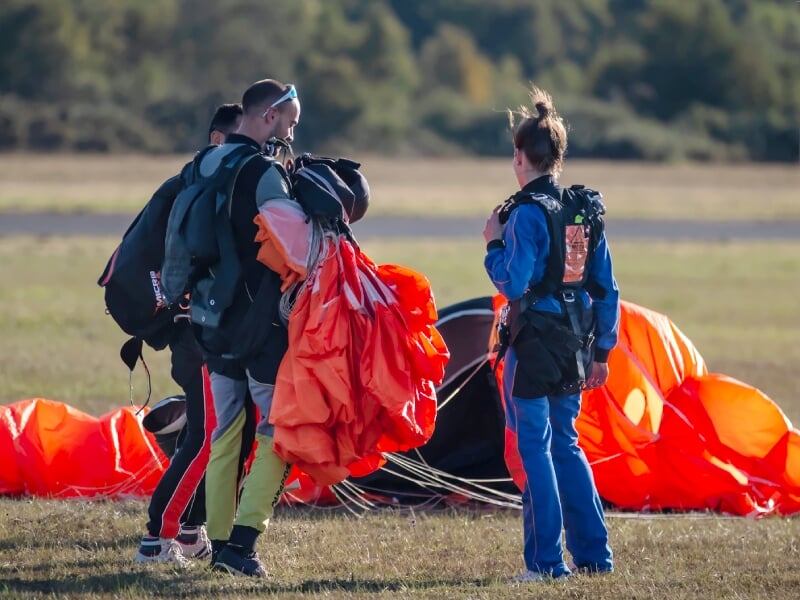 Parachutistes après atterrissage, deux hommes et une femme, équipement de saut sur le dos, voiles d'atterrissage orange et blancs recueillis, en conversation, champ herbeux à l'arrière-plan, lumière d