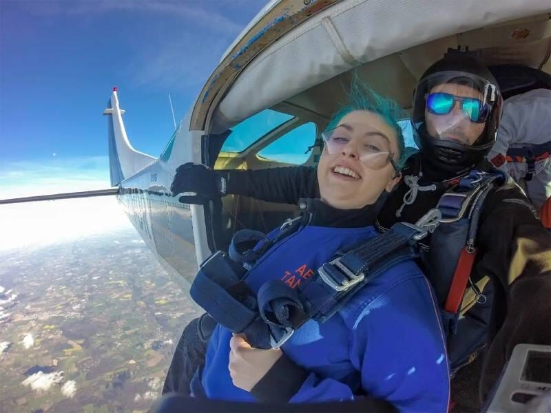 Deux personnes à la porte d'un avion en vol, prêtes à sauter dans le vide. Une femme avec le cheveux bleus et un homme avec des lunettes et un casque.
