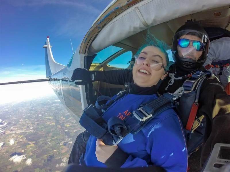 Deux personnes à la porte d'un avion en vol, prêtes à sauter dans le vide. Une femme avec le cheveux bleus et un homme avec des lunettes et un casque.