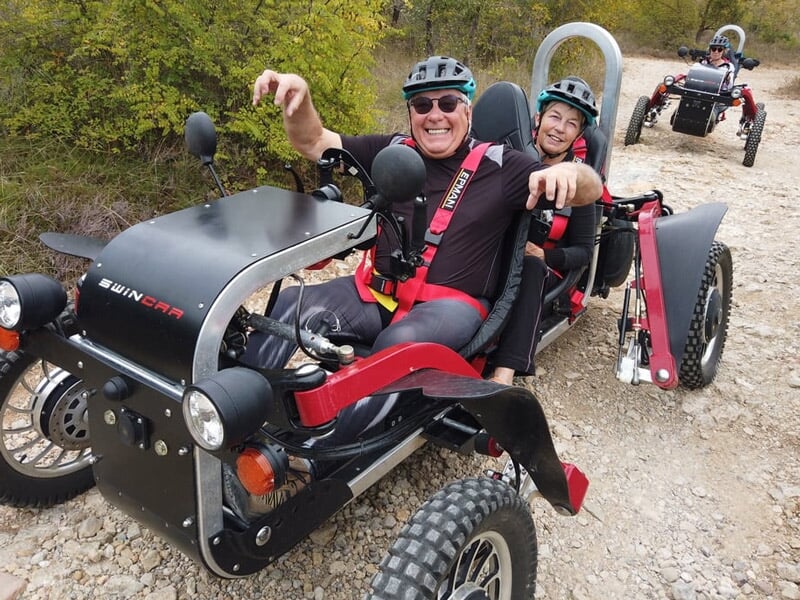 Deux personnes souriantes en tenue de sécurité sur un véhicule tout-terrain rouge et noir en chemin de terre.