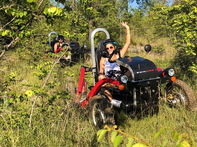 Femme souriante sur un véhicule tout-terrain rouge et noir dans une zone herbeuse avec des arbres verts.
