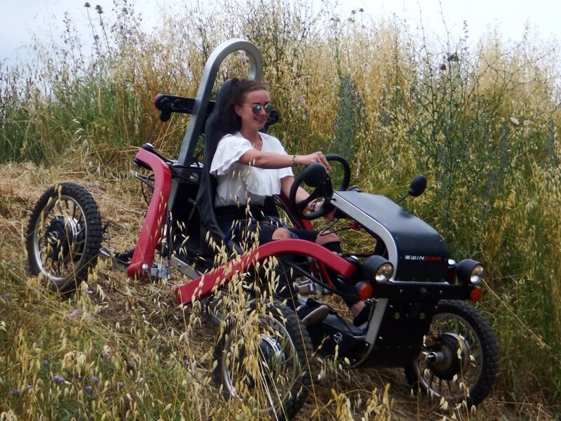 Femme au volant d'un véhicule tout-terrain rouge et noir roulant dans un champ d'herbes hautes.