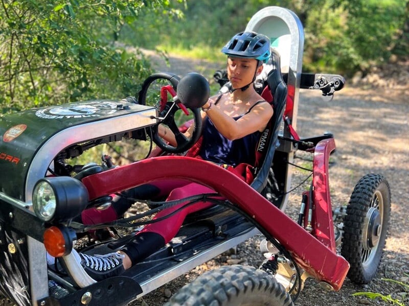 Jeune femme concentrée au volant d'un véhicule tout-terrain rouge et noir dans un environnement boisé.