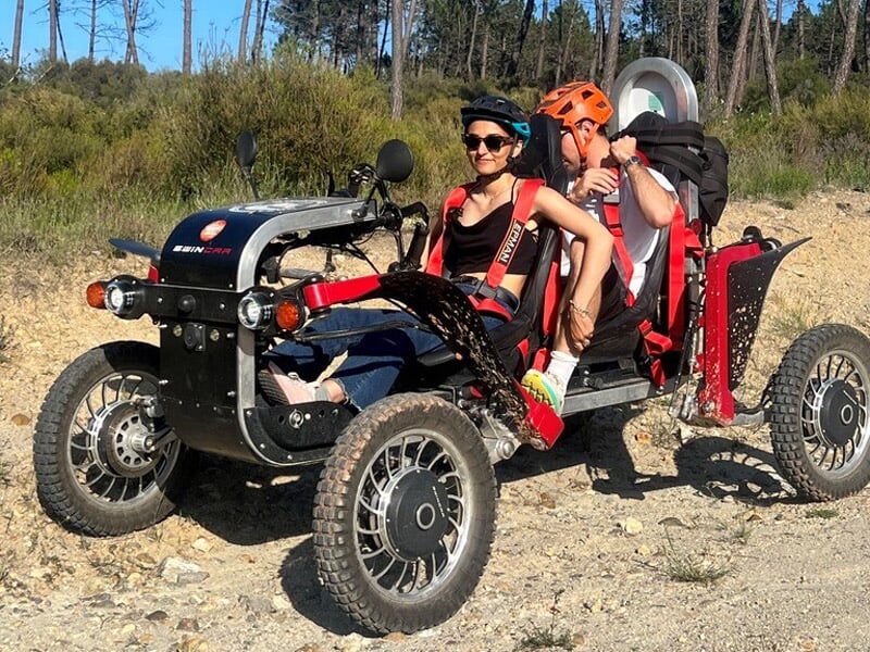 Deux personnes sur un véhicule tout-terrain rouge et noir sur un chemin de terre avec des arbres en arrière-plan.