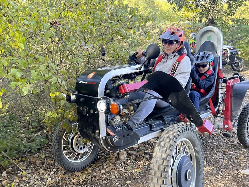 Femme et enfant conduisant un véhicule tout-terrain électrique à travers un chemin étroit entouré de feuillage.