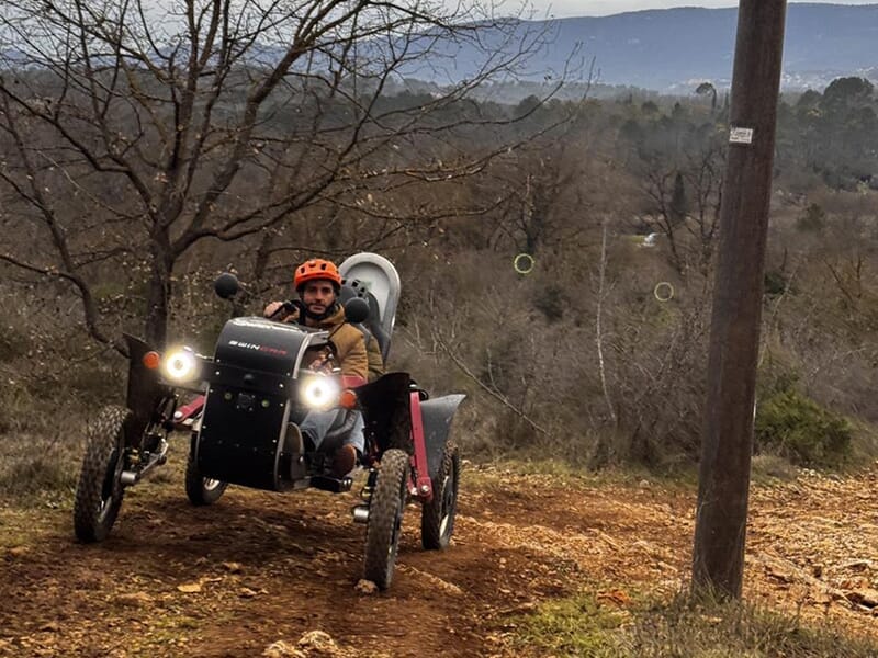 Homme seul conduisant un véhicule tout-terrain électrique en descente sur un sentier de terre, avec un paysage boisé à l’arrière-plan.