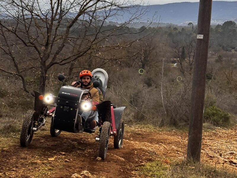 Homme seul conduisant un véhicule tout-terrain électrique en descente sur un sentier de terre, avec un paysage boisé à l’arrière-plan.