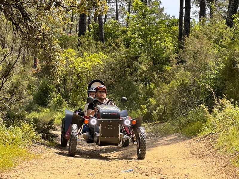Deux personnes conduisant un véhicule tout-terrain électrique sur un sentier forestier bordé de végétation dense.