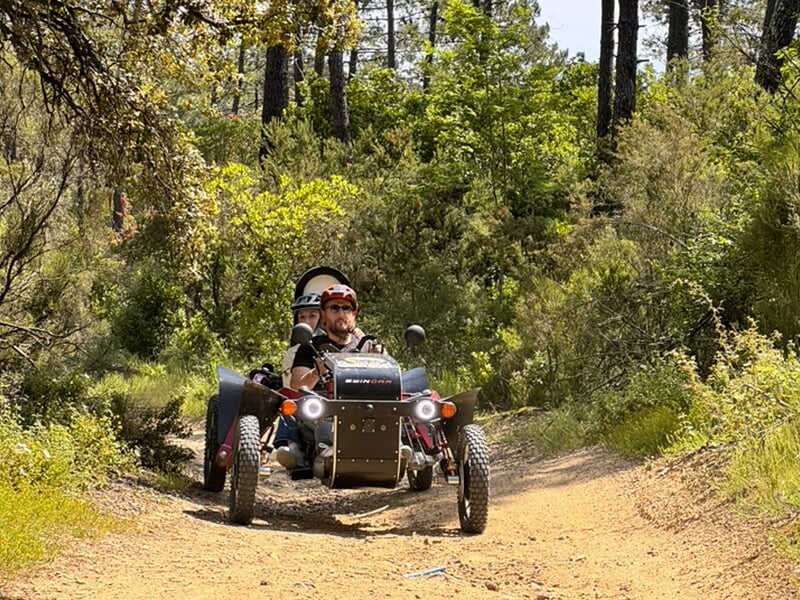 Deux personnes conduisant un véhicule tout-terrain électrique sur un sentier forestier bordé de végétation dense.