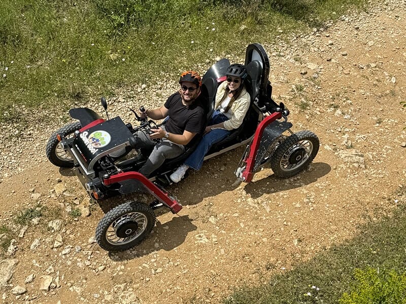 Vue aérienne d’un couple souriant dans un véhicule tout-terrain électrique sur un chemin rocailleux.