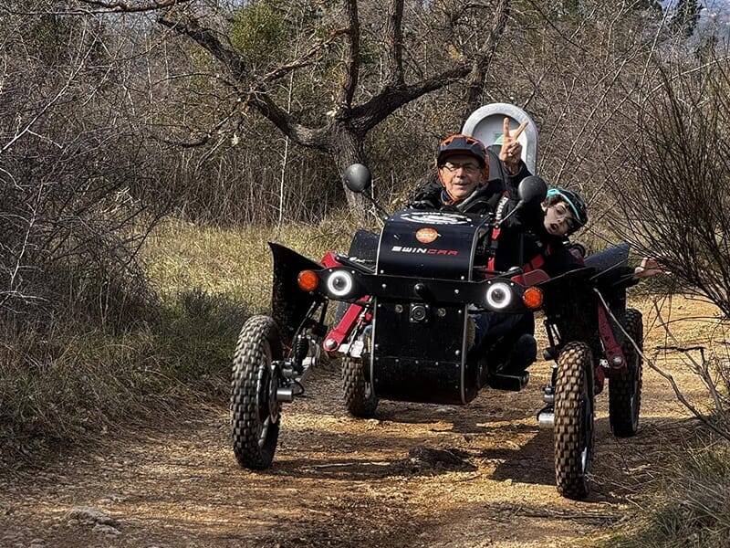 Homme faisant un signe de victoire avec les doigts en conduisant un véhicule tout-terrain électrique sur un chemin de forêt, avec un enfant passager qui sourit