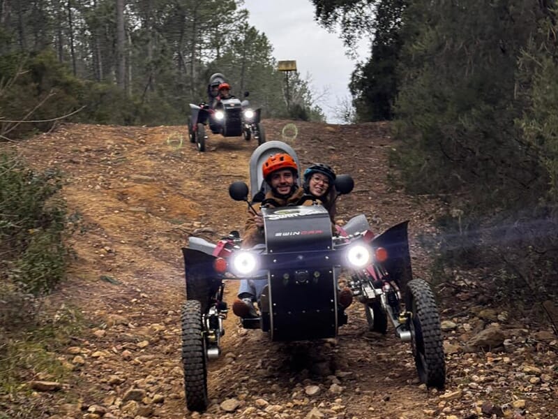 Deux personnes souriantes dans un véhicule tout-terrain électrique descendant un sentier caillouteux, suivies par un autre véhicule en arrière-plan dans un paysage forestier.