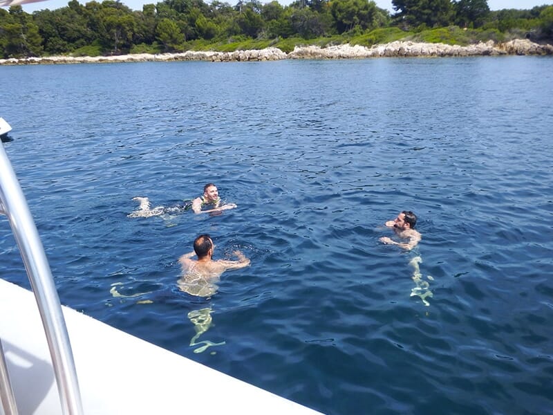 Trois personnes nagent dans une eau calme près d’une côte boisée, après avoir quitté un bateau.