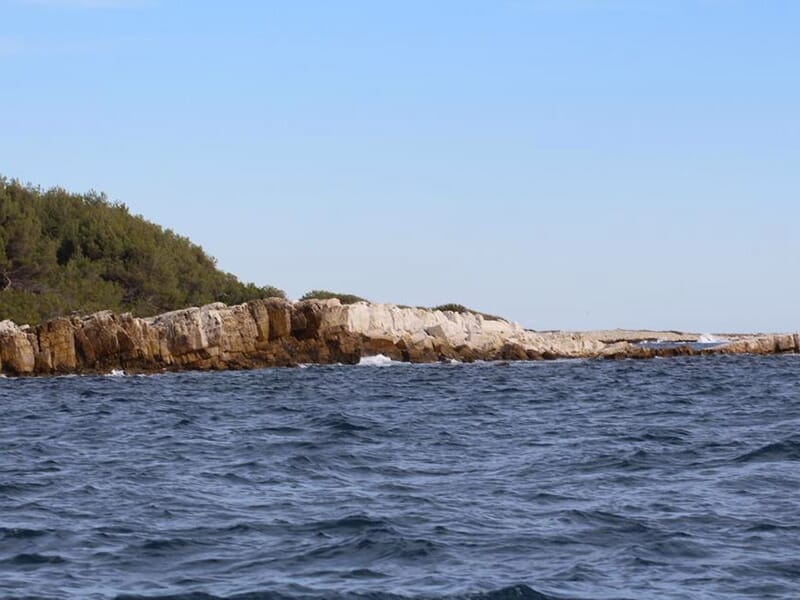 Côte rocheuse vue depuis la mer, avec des vagues venant s’écraser sur les rochers sous un ciel bleu.