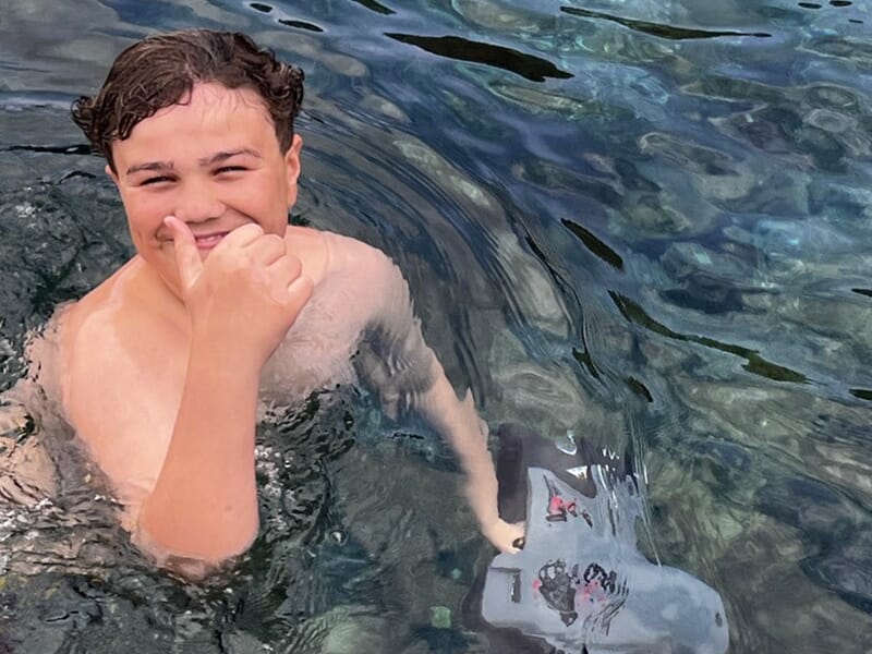 Garçon dans l’eau faisant un signe du pouce avec un grand sourire, tenant une planche de natation.