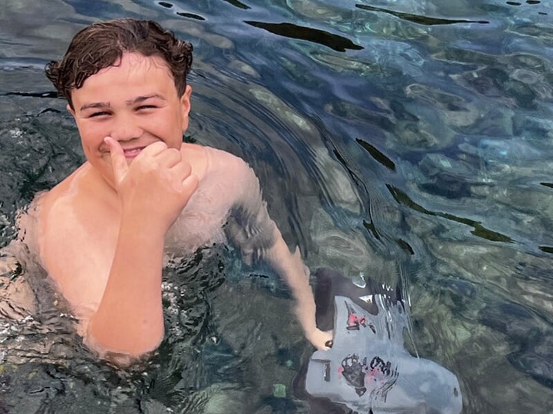 Garçon dans l’eau faisant un signe du pouce avec un grand sourire, tenant une planche de natation.
