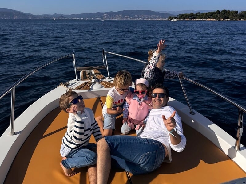 Cinq personnes, dont trois enfants, sont allongées et assises à l'avant d'un bateau en mer, souriantes sous un ciel bleu dégagé avec des montagnes en arrière-plan.