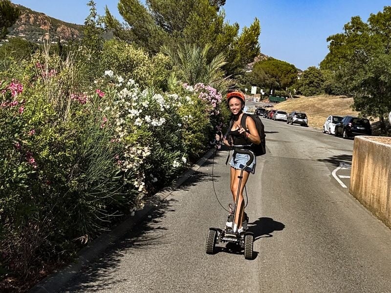 Femme souriante portant un casque roulant sur une trottinette électrique sur une route bordée de fleurs et de voitures garées.
