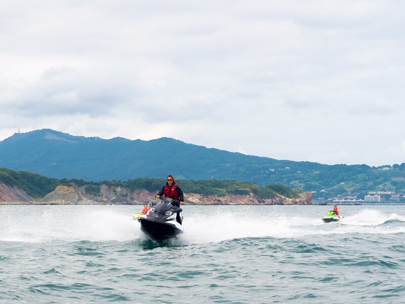 Personne conduisant un jetski noir sur une mer agitée avec des montagnes en arrière-plan sous un ciel nuageux.
