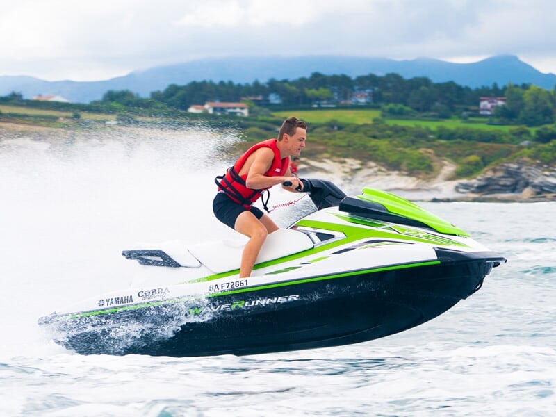 Homme en gilet rouge sautant avec un jetski vert et blanc sur une mer calme avec des collines verdoyantes en arrière-plan.