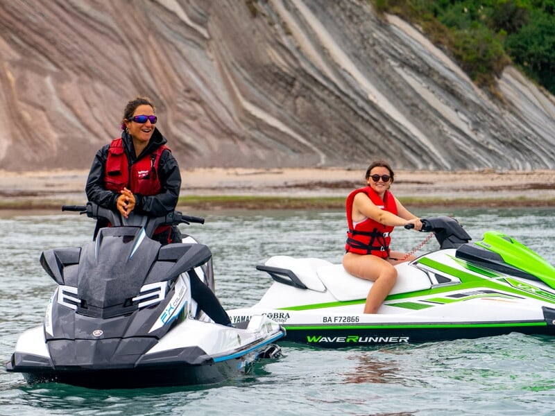 Deux personnes souriantes sur des jetskis, l'un noir et l'autre vert, près d'une plage rocheuse avec des falaises.