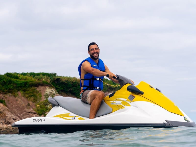 Homme souriant en gilet bleu assis sur un jetski jaune et blanc sur une mer calme avec des collines en arrière-plan.