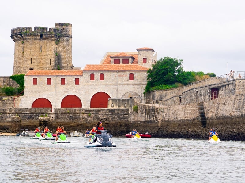 Groupe de personnes en jetskis colorés naviguant près d'un fort ancien avec des murs en pierre et des arches rouges.