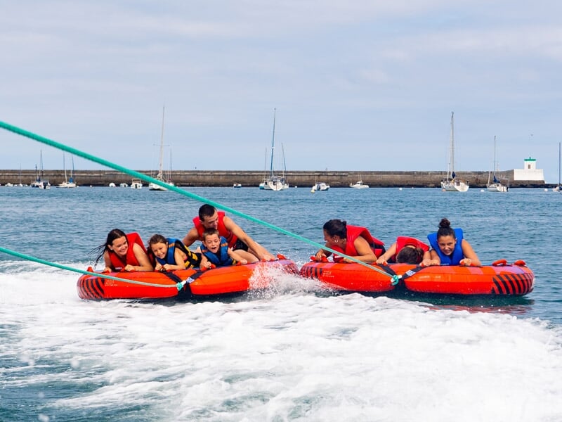 Groupe de personnes tenant des bouées tractées orange, tirées par un bateau sur une mer calme avec un port en arrière-plan.