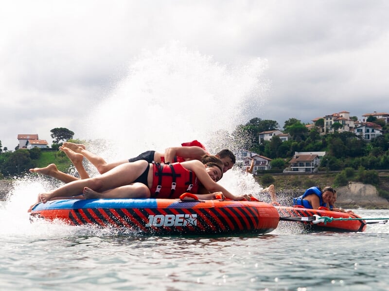 Personnes en gilets rouges et bleus sur des bouées tractées orange, éclaboussées par les vagues près d'une côte avec des maisons.