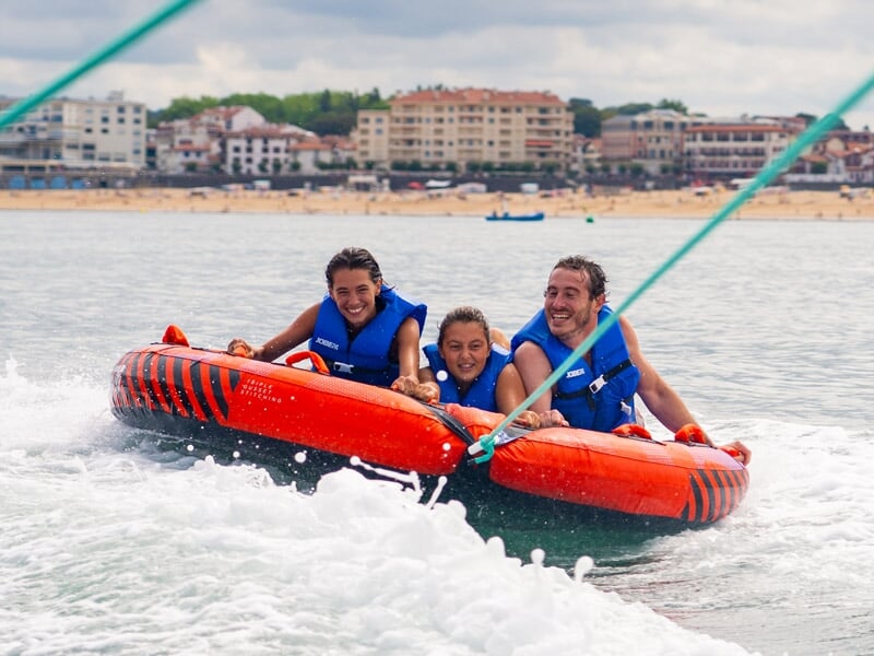 Quatre personnes souriantes sur des bouées tractées orange, tirées par un bateau sur une mer calme avec une plage en arrière-plan.