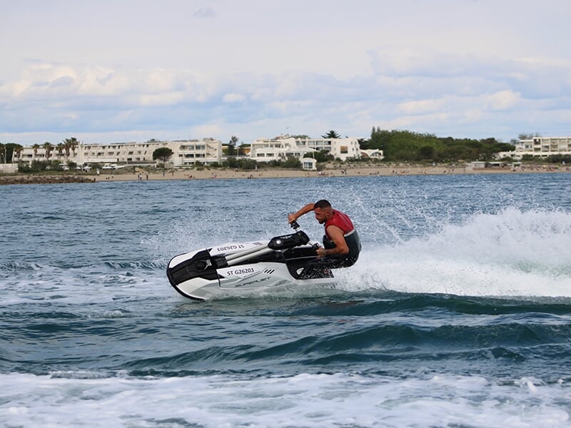 Un homme portant un gilet de sauvetage rouge et noir effectue une manœuvre sur un jet ski blanc, créant des éclaboussures sur une mer légèrement agitée, avec un littoral urbain en arrière-plan.
