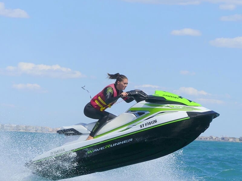 Une femme portant un gilet de sauvetage rouge et noir pilote un jet ski Yamaha WaveRunner vert et blanc, sautant au-dessus de l'eau sous un ciel bleu.