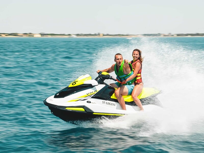 Un homme et une femme, tous deux portant des gilets de sauvetage colorés, chevauchent un jet ski jaune et blanc sur une mer calme, l'homme pointant vers l'objectif avec enthousiasme.