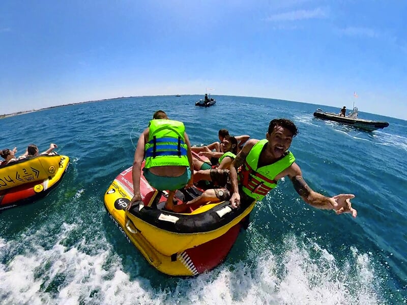 Vue arrière de deux grandes bouées gonflables jaunes tractées à pleine vitesse par un bateau, avec plusieurs passagers levant les bras en l’air sous un ciel bleu éclatant.