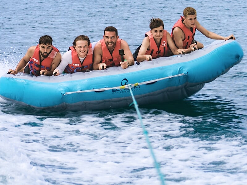 Cinq jeunes hommes portant des gilets de sauvetage rouges sont allongés sur une bouée gonflable bleue, tractée à grande vitesse sur l’eau, affichant des expressions de joie et d’excitation.