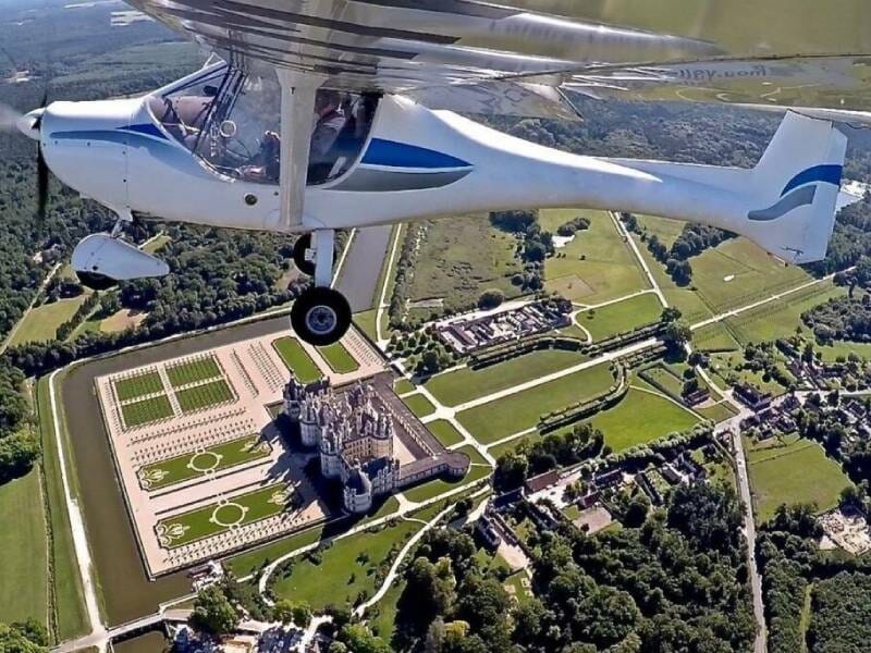Vue aérienne d'un petit avion blanc survolant un château entouré de jardins et de forêts verdoyantes lors d'un baptême en ULM près de Blois.