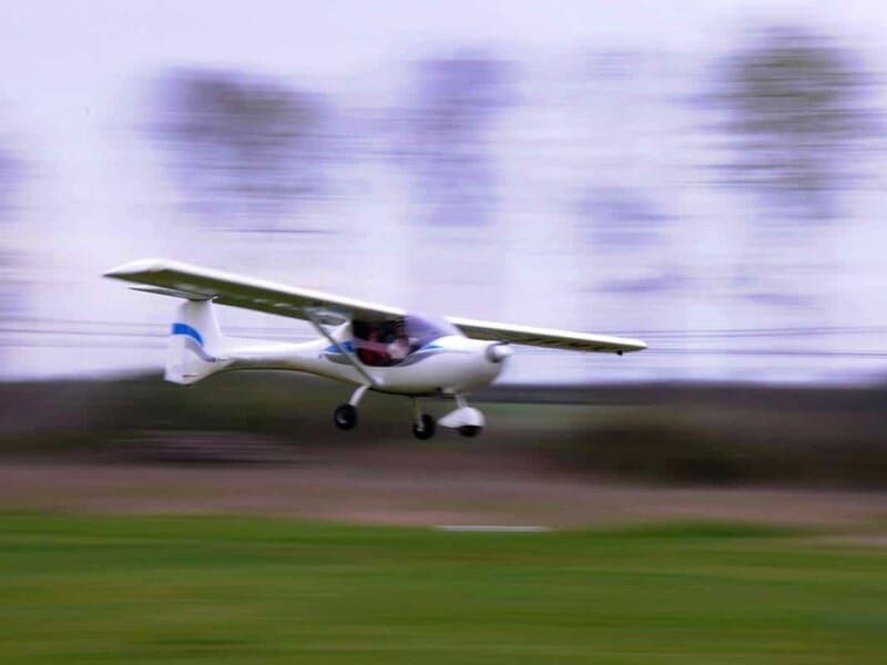 Petit avion ultraléger en vol à basse altitude au-dessus d'un champ vert avec un arrière-plan flou d'arbres.