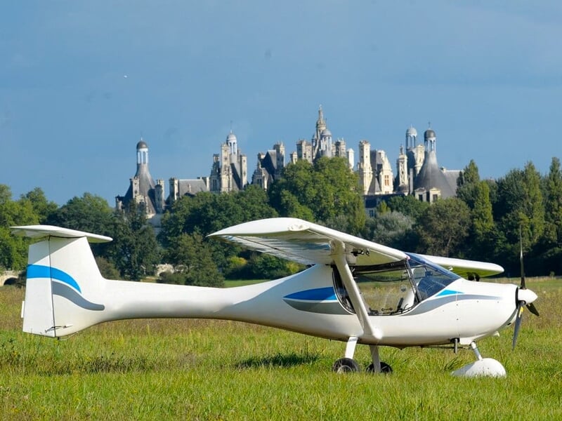 Avion ultraléger blanc et bleu stationné sur un champ vert avec un château historique visible en arrière-plan.