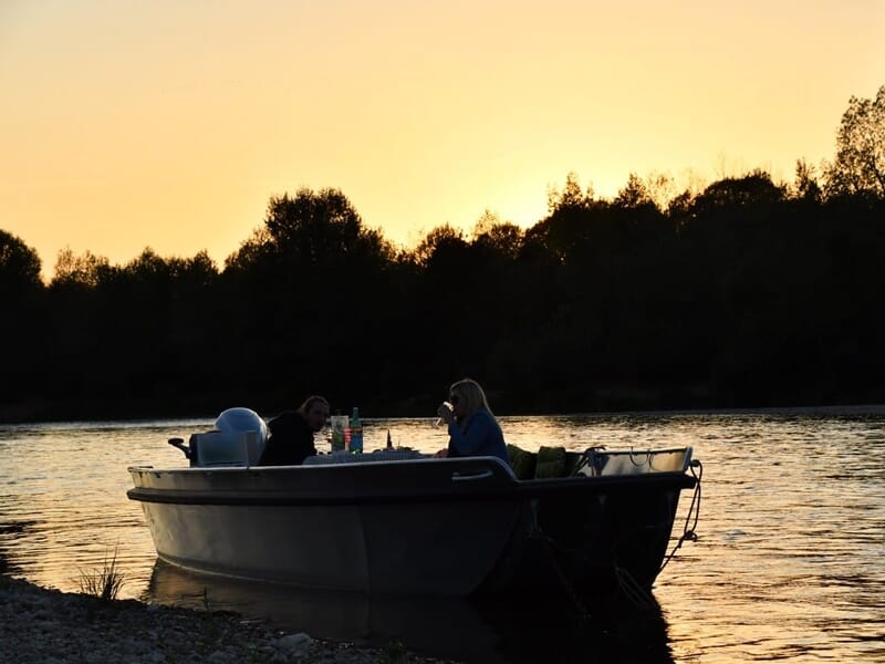 Deux personnes assises dans un bateau au bord de l'eau au coucher du soleil, ambiance calme et paisible.