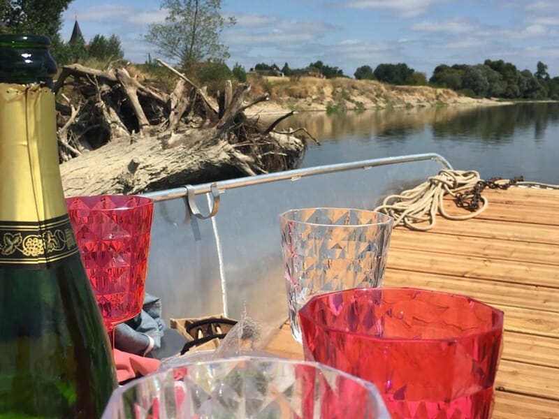 Bouteille de champagne et verres colorés posés sur un bateau avec vue sur la rivière et la rive ensoleillée.