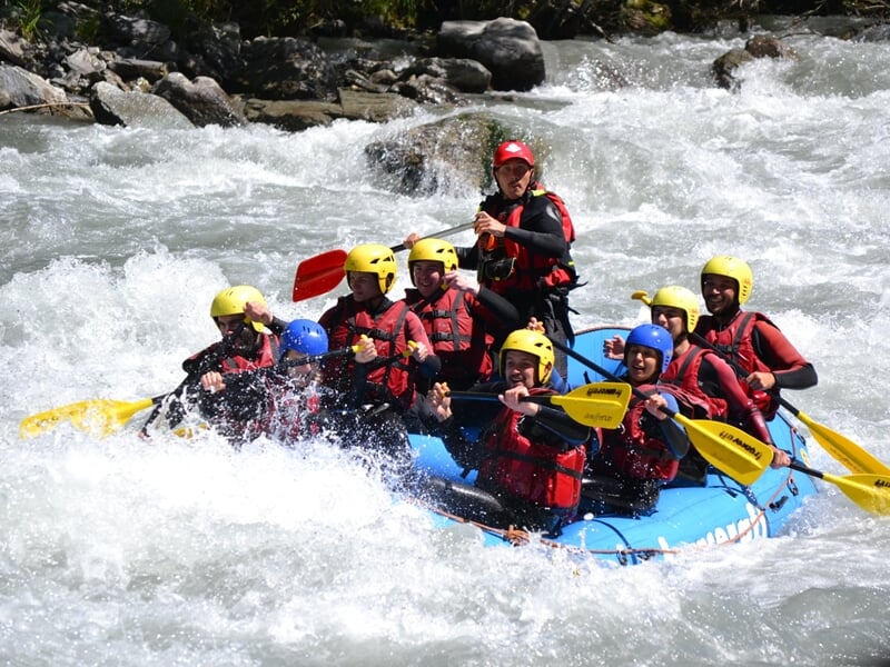 Équipe de six personnes en rafting bleu pagayant sur une rivière calme entourée de verdure.
