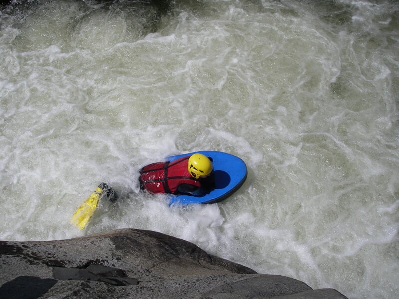 Personne en hydrospeed descend une rivière agitée, équipée d'un casque jaune et d'un gilet de sauvetage rouge.