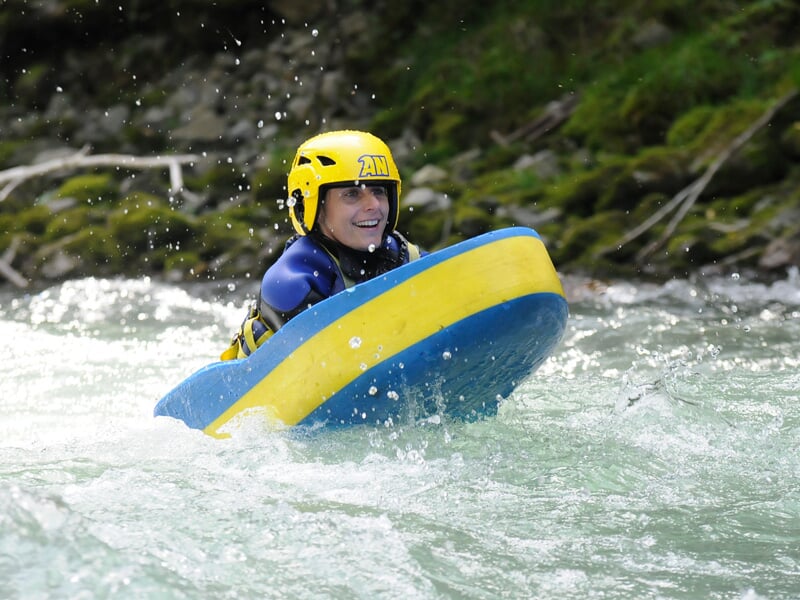 Jeune femme souriante en hydrospeed sur une rivière calme, portant un casque jaune et un gilet de sauvetage bleu.