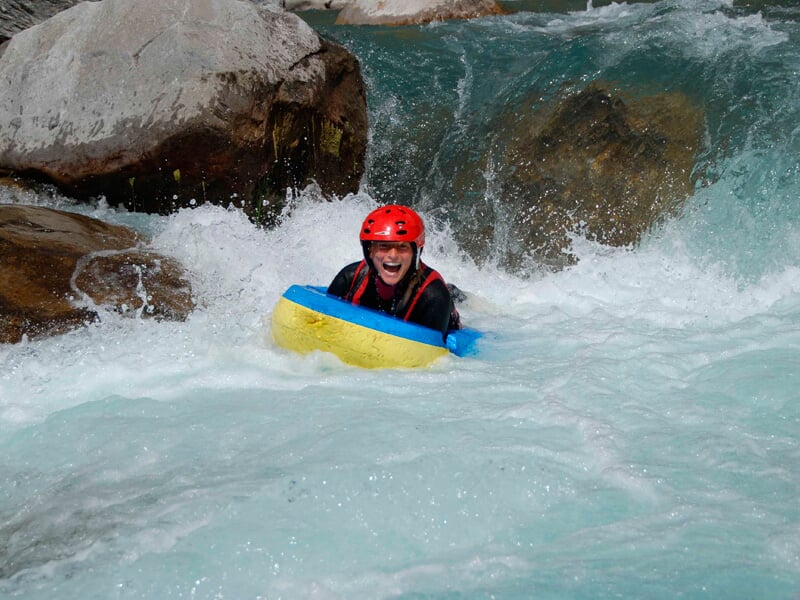 Personne en hydrospeed descend une rivière tumultueuse avec un casque rouge et un gilet de sauvetage noir.