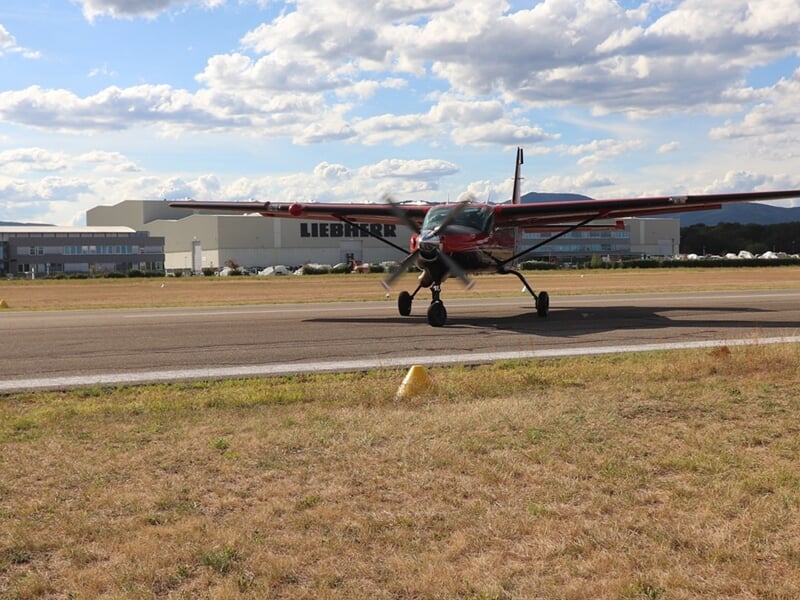 Petit avion rouge au sol sur une piste d'atterrissage entourée d'herbe sèche sous un ciel bleu.