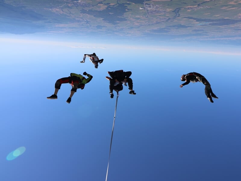 Quatre parachutistes en chute libre dans un ciel bleu clair, attachés ensemble par une corde.