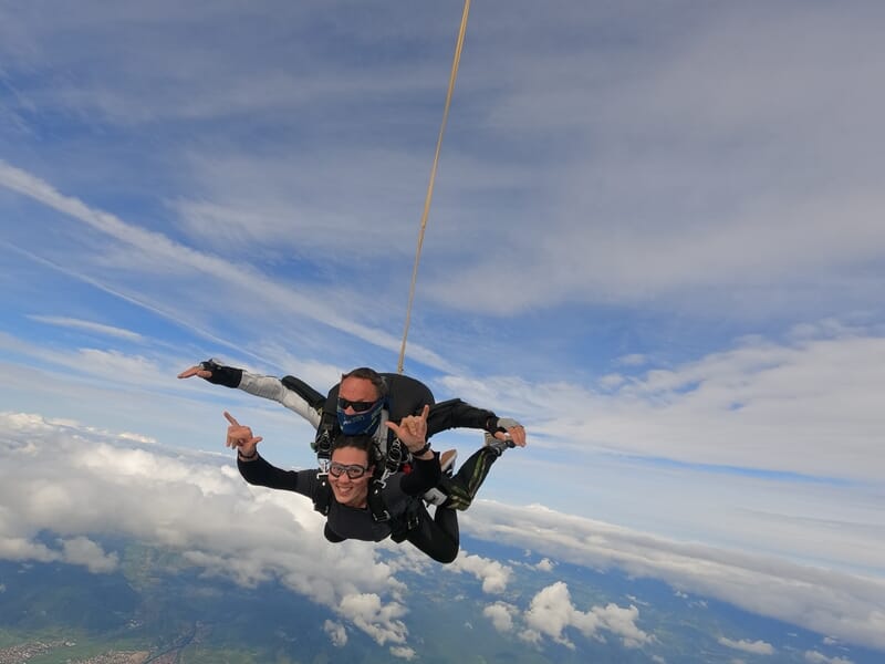 Deux personnes en parachute en tandem en plein saut, ciel bleu avec quelques nuages, expressions joyeuses.